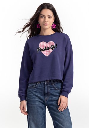 Kvinde med langt mørkt hår har en mørkeblå sweatshirt på med et lyserødt ternet hjerte og teksten "Pookie Girl", stjerneformede lyserøde øreringe og blå jeans.