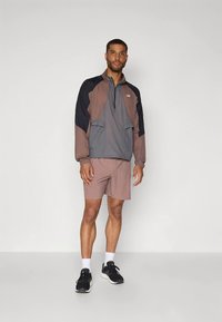 Coupe-vent gris avec des panneaux marron et bleu marine, doté d'une fermeture éclair quart, deux poches avant. Associé à un short rose et des baskets noires.