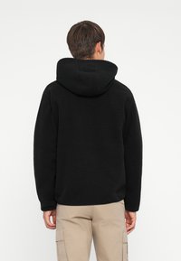 Haut en polaire noir avec une surface texturée et une grande capuche. Le vêtement a une coupe décontractée et des coutures plates, associé à un pantalon cargo beige.