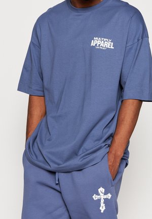 Środkowa część osoby ubranej w niebieski casualowy strój z napisem "MULTIPLY APPAREL 'LOVE PROJECT'" na koszulce oraz białym krzyżem na spodniach.