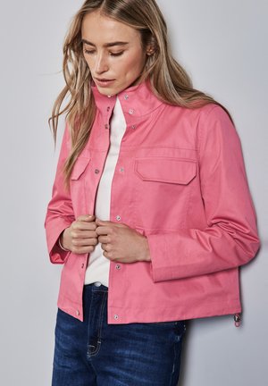 Donna che indossa un giaccone rosa con bottoni sopra una maglietta bianca e jeans blu, che guarda in basso con le mani vicino ai bottoni del giaccone.