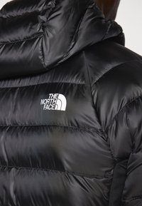 Schwarze Daunenjacke mit einem glänzenden, gesteppten Muster und einer Kapuze. Auf der oberen linken Seite befindet sich ein weißes Logo. Der strukturierte Stoff wirkt leicht.