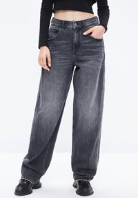 Jeans de pierna ancha en denim gris oscuro con un acabado ligeramente deslavado, que cuentan con bolsillos frontales y un cierre de botón. Llevados con calzado negro.