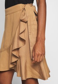 Tannfarvet slå-om-nederdel med flæsekant og sløjfedetalje ved taljen. Stoffet har en glat tekstur og en blød glans.