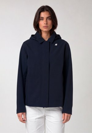 Donna che indossa una giacca con cappuccio blu navy con un piccolo logo bianco sul petto e pantaloni bianchi, in piedi contro uno sfondo chiaro e uniforme.