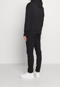 Svart huvtröja med känguruficka, svarta slim fit-byxor och vita sneakers, sedd bakifrån mot en enfärgad bakgrund.