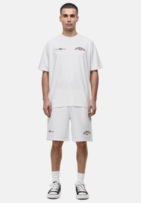 Witte katoenen t-shirt en short set, met bedrukte logo's in zwart en rood. Ruime pasvorm, ronde halslijn en platte gebreide manchetten.