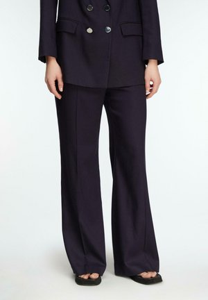 Osoba ubrana w ciemne, dopasowane spodnie i pasującą marynarkę, stojąca boso na jednolitym białym tle.