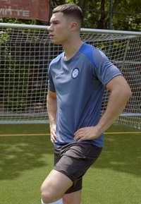 Camiseta atlética azul de mangas cortas, con logo en el pecho, combinada con pantalones cortos oscuros. Campo de hierba y porterías en el fondo.
