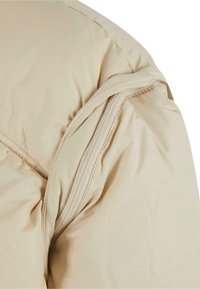 Beige puffer jacket close-up showing a zippered sleeve with gathered fabric texture and a smooth, matte finish.