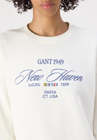Sweatshirt de couleur crème avec un texte brodé en bleu : "GANT 1949", "New Haven", "Sailing Crew" et "06516 CT. USA."