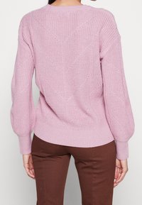 Rosa stickad tröja med ett strukturerat diamantmönster, långa ärmar med ribbade muddar och en ribbad nederkant. Bärs med bruna byxor.