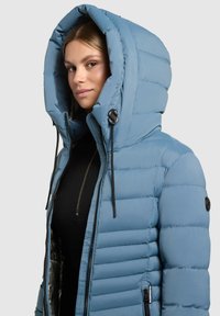 Giacca puffer azzurro chiaro con un grande cappuccio, zip frontale e dettagli in hardware nero. Presenta una texture trapuntata e lacci regolabili.