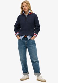 Marineblauwe gebreide pullover met rood en wit gestreepte manchetten, gedragen over een lichtblauw shirt. Jeans en crèmekleurige sneakers maken de outfit compleet.
