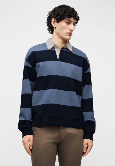 Homme portant un maillot de rugby à manches longues avec de larges rayures horizontales bleu marine et bleu, un col beige et un pantalon marron, regardant vers la droite.
