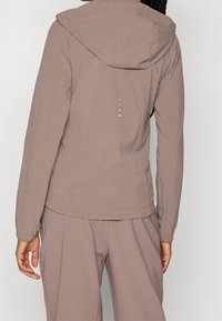 Chaqueta con capucha de color marrón claro con una textura suave y puños elásticos. Presenta detalles reflectantes y un diseño minimalista. Se muestra la vista trasera.