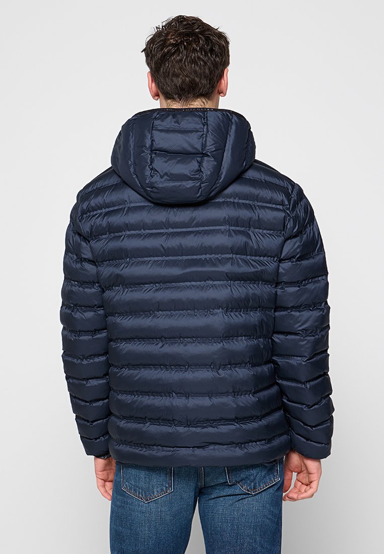 Granatowa kurtka puchowa z kapturem, z poziomymi pikowanymi sekcjami i gładką, błyszczącą fakturą. Minimalne akcenty; zaprezentowano widok z tyłu.
