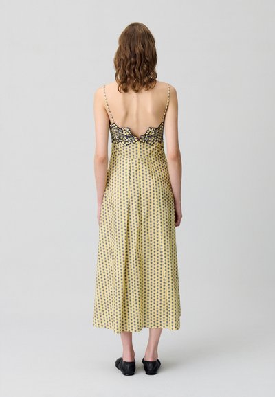 Abito giallo a fantasia con un retro in pizzo nero, bretelline sottili e una silhouette fluida che arriva sotto il ginocchio. Indossate scarpe nere.