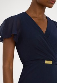 Φόρεμα ναυτικού μπλε με μανίκια φτερού, σχεδιασμό τύπου wrap και υφασμάτινη υφή. Διαθέτει χρυσή μεταλλική λεπτομέρεια στη μέση.