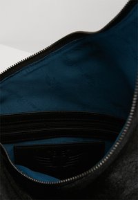 Musta nahkalaukku, jossa sileä pinta, sininen kangasvuori ja vetoketjullinen tasku. Merkittävä logo on painesetelty paneeliin.