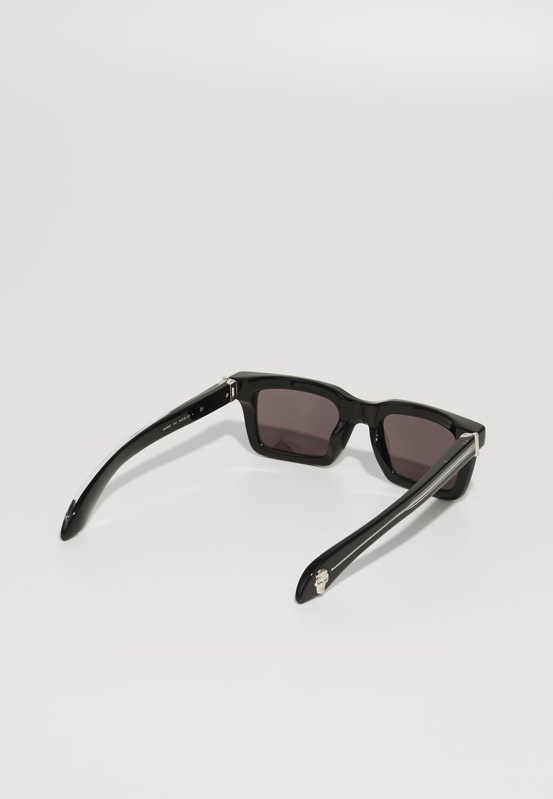 Lunettes de soleil en plastique noir avec des verres sombres, monture carrée et accents argentés sur les branches. Texture lisse et design contemporain.