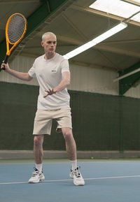 Tennise mängija valges lühikeste varrukatega särgis ja heledates lühikestes pükstes hoiab oranžide tennisereketit, seistes sinisel väljakul roheline taust.