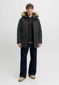 Grå parka med pelsforet hætte, store frontlommer og lynlås, båret over en mørk stribet skjorte og blå jeans med brede ben.