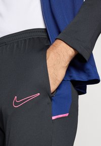Μαύρα αθλητικά σορτσάκια με ροζ λογότυπο Nike στην αριστερή πλευρά, συνδυασμένα με ναυτικό μπλε μπουφά ν που είναι ορατό στον ώμο. Λείο ύφασμα.