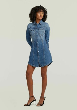 Donna con capelli ricci che indossa un vestito di jeans blu con bottoni e sandali neri con tacco alto, in piedi con le mani dietro la schiena.