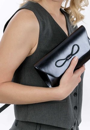 Pochette en cuir noir à finition brillante, ornée d'un nœud texturé sur le devant, tenue contre un gilet gris à décolleté en V.