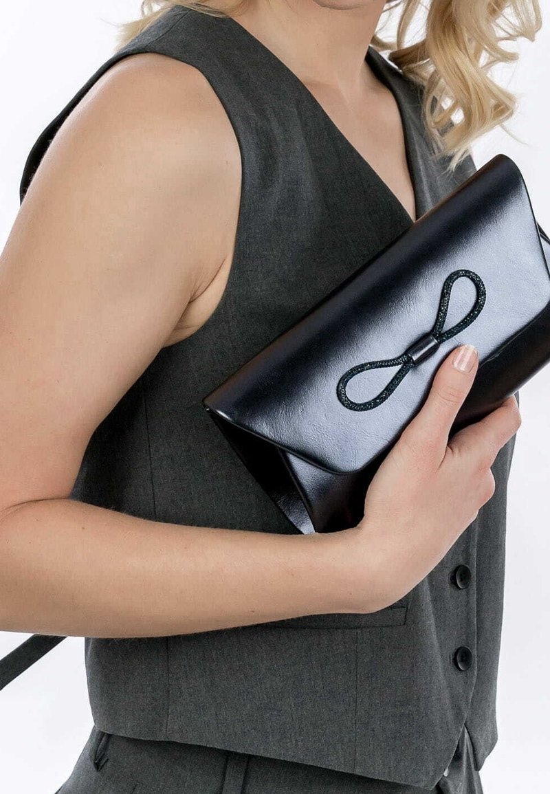 Pochette en cuir noir à finition brillante, ornée d'un nœud texturé sur le devant, tenue contre un gilet gris à décolleté en V.