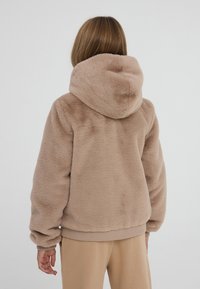 Felpa marrone e morbida con cappuccio grande e polsini a coste; texture soffice e vestibilità casual. Indossata con pantaloni beige abbinati.
