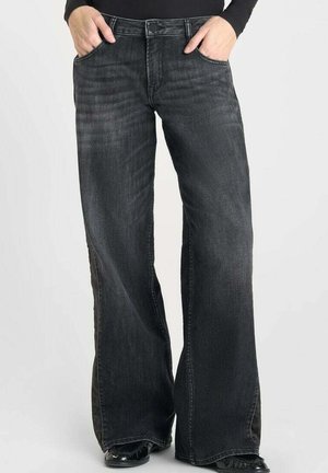 Personne portant un jean large et décontracté noir délavé avec les mains dans les poches avant, associé à des chaussures noires et un haut noir à manches longues.
