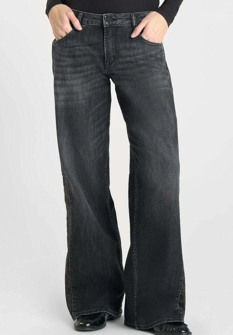 Persoon die losse, wijduitlopende, vervaagde zwarte jeans draagt met handen in de voorzakken, gecombineerd met zwarte schoenen en een zwart langemouwtop.