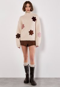 Crème turtleneck sweater met meerkleurige bloemenapplicaties, gecombineerd met bruine suède shorts en donkerbruine kniehoogte laarzen.