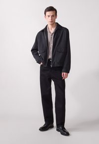 Chaqueta negra con cremallera y dos bolsillos frontales, llevada sobre una camisa de botones gris claro. Un pantalón negro y zapatos de vestir negros completan el atuendo.