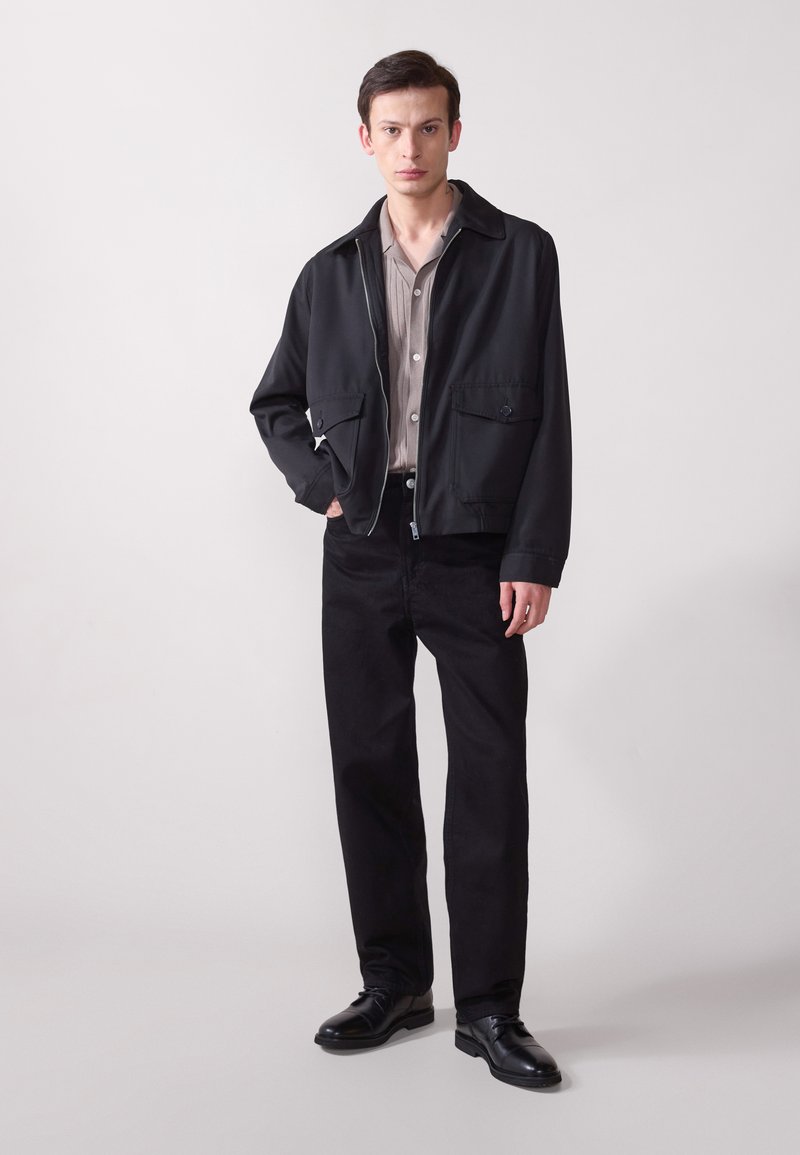Chaqueta negra con cremallera y dos bolsillos frontales, llevada sobre una camisa de botones gris claro. Un pantalón negro y zapatos de vestir negros completan el atuendo.