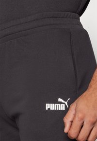 Juodos medvilnės kelnės su šoninėmis kišenėmis, elastinga juosmens linija ir balta Puma logotipu ant kairiojo šlaunies. Lygus paviršius, prilipęs dizainas.