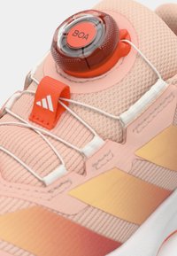 Svetlo roza športni čevelj z teksturiranim zgornjim delom, BOA gumbom za nastavljivo zavezovanje in oranžnimi poudarki na jezičku ter stranskih črtah.