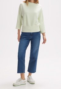 Femme portant un pull côtelé vert clair, un jean court bleu aux ourlets effilochés, et des baskets blanches à semelles vertes.