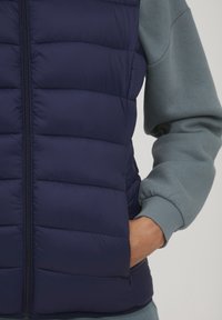 Marine blauwe puffer vest met een gewatteerd ontwerp, rits aan de voorkant en zijzakken, gelaagd over een licht turquoise sweatshirt met geribde manchetten.