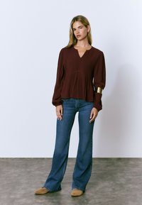 Blusa burdeos de cuello en V con mangas abullonadas, tela texturizada, cintura fruncida, combinada con vaqueros azul de campana y zapatos de lona en tono beige.