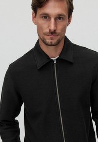 Sort fleecejakke med lynlås, ståkrave og struktureret stof, der har en slim fit og minimalistisk design.