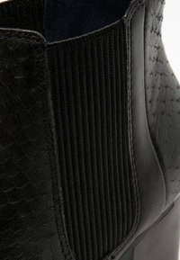 Gros plan d'une botte cheville en cuir noir avec un panneau latéral élastique côtelé et un motif texturé sur la surface du cuir.
