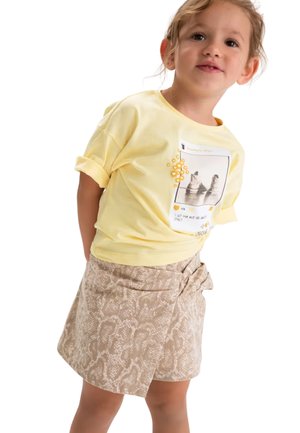 Bambino piccolo con t-shirt gialla con stampa grafica e gonna beige con stampa a serpente e fiocco, in piedi davanti a uno sfondo bianco.