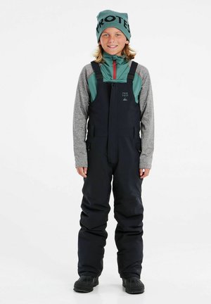 Salopette nera con bretelle regolabili, tasche con zip e design impermeabile, abbinata a una maglietta a maniche lunghe teal e grigia e un berretto.
