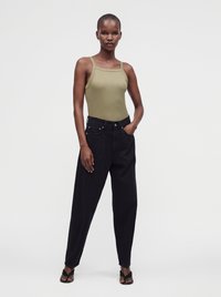 Femme debout avec une main dans la poche, portant un haut sans manches vert olive, un pantalon noir ample et des sandales noires à bouts ouverts sur fond blanc.