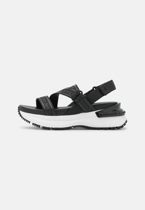 Sandales sportives noires avec plusieurs brides réglables, une semelle intermédiaire blanche épaisse, une semelle extérieure noire texturée et des détails de branding subtils.