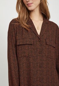 Brun mönstrad blus med krage, två bröstfickor och ett mjukt, strukturerat tyg med abstrakta orangeprickar.