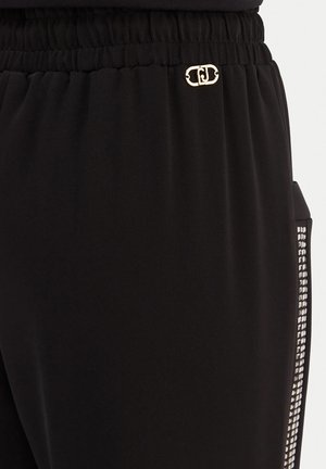 Mustad püksid elastse vööümbrisega, väike kuldne logo vöö lähedal ning hõbedastest ruudukujulistest naastudest vertikaalne rida küljetaskus.